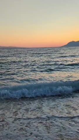 The video shows a peaceful beach scene at sunset. The sun is setting over the ocean, casting a warm orange and yellow glow across the sky. Gentle waves are rolling onto the shore, creating a soft, calming rhythm. The beach is sandy and appears to be empty. The overall atmosphere is one of tranquility and natural beauty.