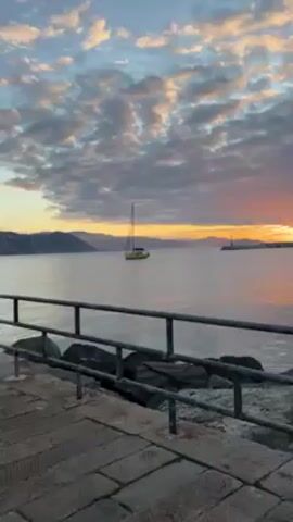 The video showcases a serene coastal scene at dawn or dusk. The camera pans across a stone pier, revealing a metal railing along the water's edge. In the distance, a green and yellow sailboat rests on the calm sea, framed by a backdrop of hills and a sky filled with soft, pastel-colored clouds. As the camera moves, a row of seagulls perched on the railing comes into view. The overall setting evokes a sense of tranquility and natural beauty.