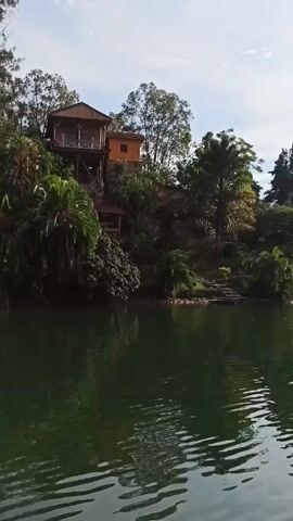 The video pans across a green lake towards a hillside with lush vegetation. Several houses are built on the hillside, including wooden structures elevated on stilts. Two beach umbrellas are visible on a grassy area near the water's edge. A person's voice mentions the sunset.