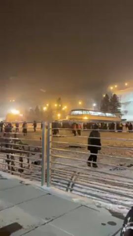 The video depicts a nighttime scene at a ski resort. People are seen on a snow-covered slope with ski lifts visible in the background. The area is lit by artificial lights, and snow is falling. A person in dark clothing walks away from a metal railing toward the ski slope. Ski poles and skis are placed on the snow near the railing. The scene is somewhat hazy, with the background appearing slightly blurred due to the falling snow.