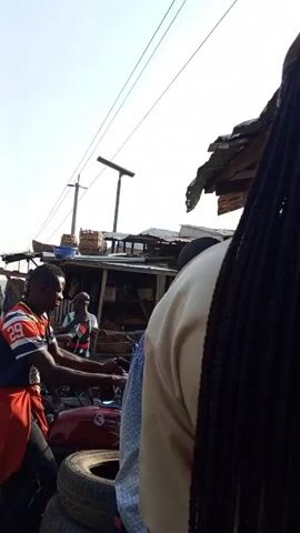 The video shows a bustling outdoor market in what appears to be a developing country. Several people are visible, many of whom are wearing traditional or modest clothing. There is a woman in a hijab carrying a child on her back, while another is wearing a headscarf. Several men are present, some are wearing Western-style clothing such as T-shirts and pants, while others are in more traditional garments. Several motorcycles are parked on the side of a dirt road, some of which are stacked with tires. The market stalls are built with basic materials like wood and corrugated metal. Power lines stretch across the sky overhead. The overall scene suggests a vibrant, crowded marketplace with a mix of traditional and modern elements.