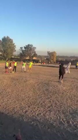 The video shows a group of people playing soccer on a dirt field on a sunny day. Some of the players are wearing neon green or yellow vests over their clothing. One person on the sideline is wearing all black clothing. In the background, there are trees and a fence. Some of the players are kicking a soccer ball. There is a small soccer goal on the left side of the frame. The video appears to have been taken at ground level.