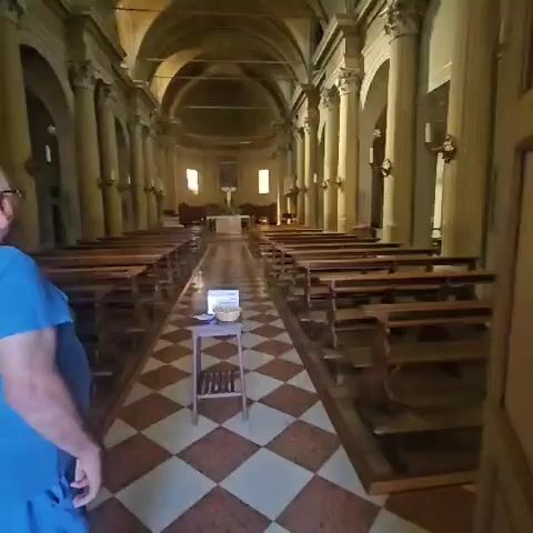 The video shows the interior of a church. The camera pans slowly down the central aisle, which is flanked by rows of wooden pews on either side. The floor is tiled in a red and white diamond pattern. At the far end of the aisle, the altar is visible, with a crucifix and a painting of Jesus above it. There is also a smaller table with a basket of items, and a sign in Italian, in the foreground. Sunlight streams in through stained glass windows, illuminating dust motes in the air. The church appears empty and quiet.