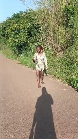 The video shows a young woman walking on a paved road. She has dark hair tied up in a ponytail, and she's wearing a patterned shirt over a white top and short denim shorts. She's also wearing flip-flops. She's holding a phone in one hand and something else in the other. The road is lined with greenery on both sides, with tall grass and bushes. The shadow of the person recording the video is visible on the road. It's a sunny day.