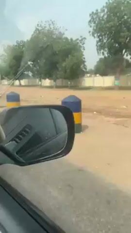 The video shows a view from inside a moving vehicle, likely a car, driving on a dirt road or street. The vehicle passes by several cylindrical bollards painted with bands of blue, yellow, and green. Beyond the bollards, there's a dirt area, a low white wall, and trees. The side view mirror of the car is visible in the foreground, and other vehicles can be seen in the reflection. The sky is light, suggesting it's daytime. The scene appears to be in a rural or suburban area.