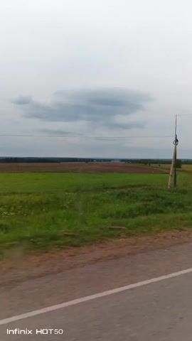 This video shows a view from a moving vehicle on a cloudy day. The scene is a rural landscape with green fields, a dirt road, and utility poles with wires. In the distance, there are trees and some structures that appear to be houses or buildings. The video is characterized by visual distortions, suggesting a technical issue with the recording or playback.