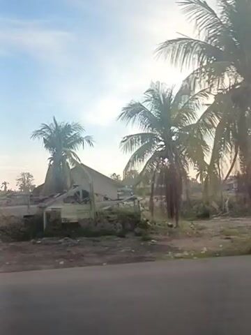 The video shows a series of scenes of what appear to be damaged or abandoned buildings and structures in what seems to be a rural or semi-rural area. In the first few frames, there is a damaged structure with a collapsed roof, possibly from a storm. Coconut trees are visible, suggesting a tropical or subtropical environment. Debris is scattered around the damaged buildings. In some frames, there are more intact but weathered buildings. One scene shows a person riding a motorcycle past a building. The sky is visible throughout, with clear blue skies and some wispy clouds, and the lighting suggests it is either early morning or late afternoon. The overall impression is one of disrepair and possibly neglect in a tropical setting.