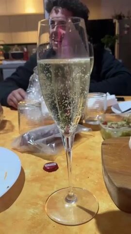 The video shows a tall glass filled with a sparkling beverage, likely champagne or Prosecco, sitting on an orange tablecloth. The bubbles in the glass are visible.  Around the glass, there is a plate, a small glass container, a package, a red bottle cap with "PERONI" printed on it, a bowl containing a green substance, and a wooden cutting board. A person is seated in the background, wearing a black jacket and glasses. The person appears to be conversing with someone. The kitchen area in the background includes cabinets, houseplants, and other kitchen items.