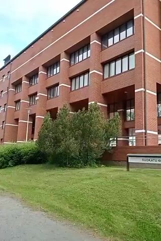 The video shows a brick building with white accents. A sign in front of the building reads "SUOKATU 44". There is a grassy area in front of the building and some trees on the left. The sky is cloudy. The camera is panning from left to right.