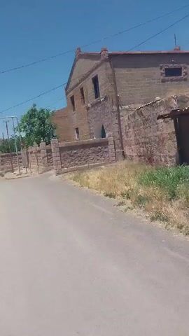 The video shows a street in what appears to be a rural or historical setting. A weathered brick building with an arched doorway and multiple windows stands on the right side of the frame. A stone wall, also constructed from weathered bricks, runs along the front of the building and extends down the street. A single car is parked on the left side of the street, near a metal gate and a utility pole. A green tree grows behind the wall, and dry grass and weeds are visible along the roadside. The sky is clear and blue, indicating sunny weather. The camera is moving, suggesting the video was captured from a vehicle or while walking.