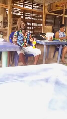 The video shows a man in a brown and black polo shirt and colorful floral shorts standing between two blue plastic tables with people sitting at them. At the first table, a man in a colorful tropical shirt and white shorts sits holding a yellow soda bottle. Another person sits at the table behind him. At the other table, a man sits with a plate of food and a brown bottle. The video is shot from a lower table covered in a blue tarp. The building is made of wood and metal sheeting.