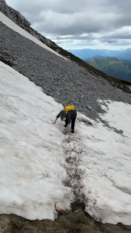 A person with a yellow backpack is climbing up a snow-covered mountain slope. The person is on all fours, using their hands and feet to navigate the snowy path. The slope is steep and rocky, with patches of snow and dirt. In the background, there are rolling green hills and mountains under a cloudy sky.