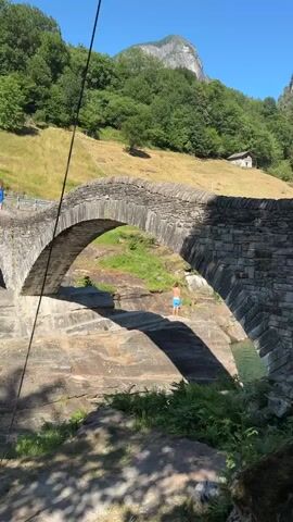A person in blue shorts stands on a rocky ledge beneath a stone bridge. They are facing away from the camera. Another person in blue is visible on the bridge, walking away from the camera. The bridge has two arches and is made of stone. There are trees and a grassy hill behind the bridge, and a mountain peak in the distance. The water beneath the bridge is visible and appears to be clear. A thin black cable or wire runs diagonally across the frame. The lighting suggests it is a sunny day.