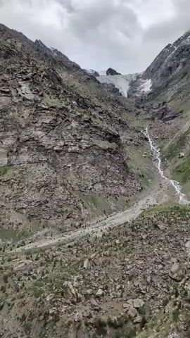 A video of a steep mountain valley with a glacier at the top. A stream runs down the side of the mountain, and there is a road with a utility pole next to it. The sky is cloudy, and the mountains are rocky and covered with sparse vegetation. The video is filmed from a moving vehicle.
