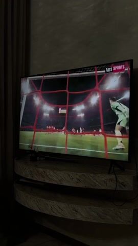 A television displaying a soccer game is situated on a wooden stand. The camera shows the game from behind the goal, with the goalie clad in green jumping in an attempt to block the ball. Several players are visible in the background. The TV screen is labeled "FAST SPORTS" in the upper right corner. The room is dimly lit, with dark curtains visible on the left side of the frame.