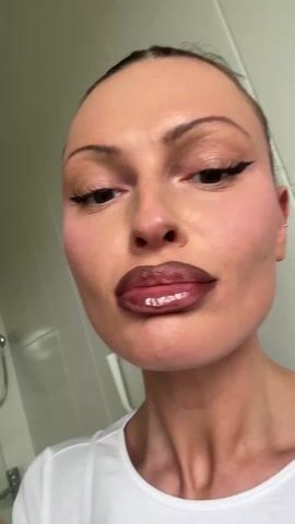 A woman with her hair pulled back and wearing winged eyeliner and a white top is shown applying a lip product. She holds a white tube with a metallic rollerball applicator to her lips, which are already made up with a dark liner and a lighter shade in the center. She moves the applicator across her lower lip, then puckers her lips, showcasing the glossy effect. Subsequently, she applies the product to her upper lip, looking directly at the camera. The lip product gives her lips a shiny, plump appearance. The background is a light-colored wall, possibly a bathroom, with a faint reflection of what looks like a shower fixture visible.