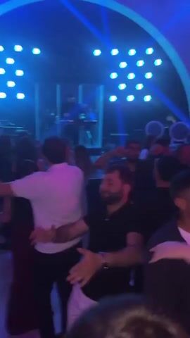 The video shows people dancing at “The Morena Hall.” Many partygoers are moving along to the music in dim, flickering lighting. A DJ stands behind a deck at the back of the room. The light periodically flashes green, white, and blue. A few people hold their arms up. The people in the foreground have various skin tones, and they seem to be enjoying the music and dance.