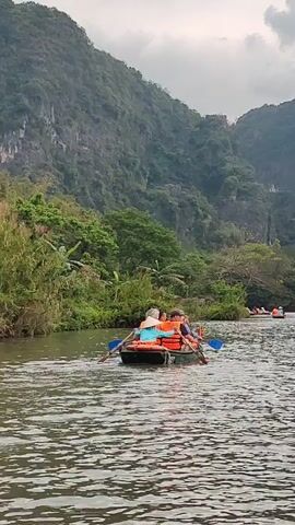 The video shows a river scene with several small boats being rowed along a river with a mountain range in the background. The sky is overcast. The boats are filled with people wearing life vests, many are rowing. Lush green vegetation lines the riverbanks and covers the mountains in the background.  The water is choppy.