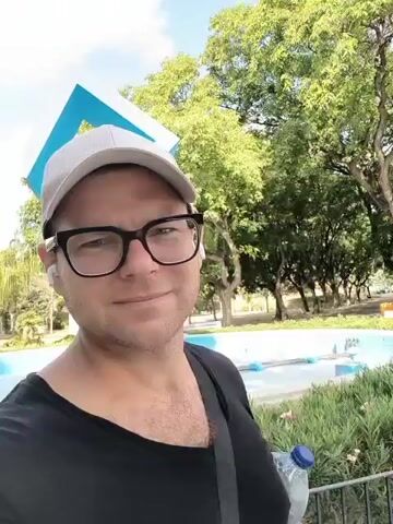 The video is a selfie-style recording of a man wearing a white baseball cap, black-rimmed glasses, and a black t-shirt. He appears to be outdoors in a park-like setting. The background shows lush green trees, palm trees, benches, a pathway, and a blue structure that might be a fountain or part of a playground. A large truck is visible in the distance behind him. The man is holding a plastic bottle of water with a blue cap in his right hand. He smiles at the camera. The audio contains the man speaking in Russian, describing the view as a "terrible vista."