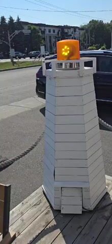 A homemade lighthouse structure, painted white, stands on a wooden surface. It has a hexagonal base and tapers upwards. The structure has horizontal planks and a small door-like opening at the bottom. A bright orange, flashing light is mounted on top. A thick rope railing is visible to the left and right of the lighthouse. In the background, a street with cars and buildings can be seen. The video is a short, static shot of the lighthouse.