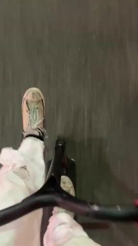 The video begins with a blurry shot of a sidewalk and the underside of what appears to be a stroller or shopping cart. It then transitions to a perspective from the street, looking down a city street at night. Buildings line both sides of the street, adorned with neon signs and lights. Cars are visible driving down the street, their headlights illuminating the wet asphalt. Pedestrians are also present on the sidewalks. The camera continues to move forward, capturing the bustling urban environment.