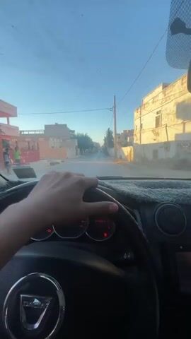 A first-person perspective video shows a hand holding the steering wheel of a Dacia car as it drives down a street lined with buildings. The sun is setting, casting long shadows. The dashboard displays the odometer reading 80984.