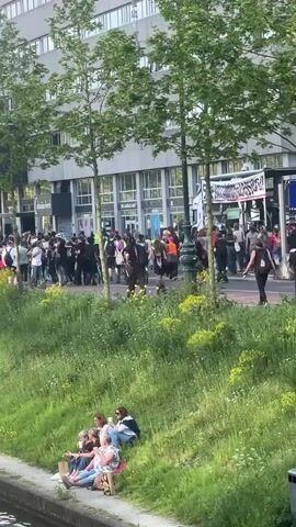The video shows a demonstration or protest taking place on a city street. A crowd of people, dressed in a variety of clothing, is gathered behind a banner, some holding signs.  Two police vans and several officers are present at the scene, suggesting a possible crowd control situation.  In the foreground, a group of five people are sitting on a grassy bank overlooking the street. The setting appears to be a city with trees and buildings lining the street.
