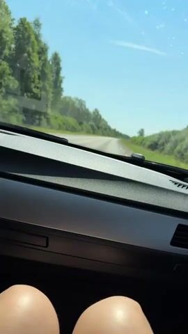 The video shows a view from inside a car. The dashboard is visible in the foreground, with a person's knees in the lower part of the frame. The windshield shows a road curving ahead, flanked by green trees and vegetation under a clear blue sky. The car appears to be moving on a sunny day.