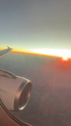 The video shows a view from an airplane window during sunset. The sun is setting on the horizon, casting a bright orange glow. The wing and engine of the airplane are visible in the foreground. The landscape below appears to be dotted with lights, possibly from a city or towns. The sky is clear and transitions from a light blue at the top to orange and yellow near the horizon.