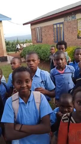 A group of African children stand outside a building in the daylight. Most of the children wear light blue shirts and some wear backpacks. The building has a sign that reads "WELCOME." Some of the children wave at the camera. One child holds a water bottle. The landscape is visible in the background.