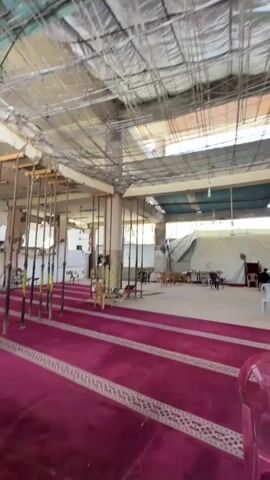 The video pans across an interior space that appears to be a place of worship, possibly a mosque, due to the presence of prayer rugs. The ceiling is exposed and shows a complex network of scaffolding and construction materials, suggesting ongoing renovation or damage repair. The lighting is somewhat dim, with natural light filtering in from large openings or windows at the far end of the space.

In the foreground, a bright pink plastic chair is prominently placed on a deep red prayer rug with a white geometric border. The rug extends throughout the visible portion of the floor.

Further into the space, several rows of prayer rugs are laid out. Along the left side, a series of tall, thin poles, possibly part of the scaffolding or used for support, are arranged in a line.

In the middle ground, several people are present, mostly seated. One individual is visible in a white chair. There are tables and chairs set up, and some people appear to be engaged in conversation or prayer. A large tent-like structure is visible in the background, further indicating an incomplete or disrupted state of the area. The walls are a neutral color, and some decorative elements, like patterned pillars, are visible. The overall impression is one of a functional space that is currently undergoing significant modification or restoration.