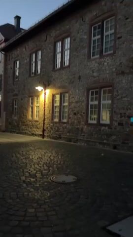 This media file depicts a nighttime street scene in Butzbach, Germany. The primary subject is a cobblestone street illuminated by a single, old-fashioned street lamp. The street lamp casts a warm, inviting glow on the textured cobblestones and the stone facade of a building on the right. This building features several large, multi-paned windows with reflective glass, suggesting it may be an older structure.

The scene is set at dusk or early night, as indicated by the dim, twilight sky visible above the buildings. The weather appears to be clear, with no precipitation. To the left of the main building, another building with a lighter facade and red-tiled roof is visible, along with some cars parked along the street and a few rose bushes. A blue waste bin is also present on the left.

There are no people visible in the frame, contributing to a sense of quiet and stillness. The overall mood is peaceful and perhaps a little mysterious, enhanced by the limited light and the absence of human activity. A circular manhole cover is prominent in the foreground on the street. A street sign with yellow and blue elements is visible on the left side of the street, partially obscured by the rose bushes.
