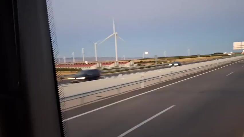 A car speeds down a highway, with wind turbines visible in the distance against a clear sky. The sun is setting, casting a warm glow on the landscape, and a large full moon is visible in the sky. The car is the only vehicle visible on the highway. The scene is peaceful and serene.