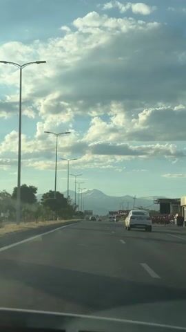 The video shows a view from a car driving on a highway. The road has multiple lanes and is bordered by a grassy median and trees on the left. Streetlights line the side of the road. In the distance, a snow-capped mountain range is visible against a sky filled with dramatic clouds. Several cars and trucks are on the highway, traveling in the same direction as the camera. The overall scene is one of travel on a sunny day with a majestic natural backdrop.