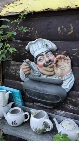 The video shows a close-up of a wall decoration of a chef holding a spoon over a pot that reads "Smells Good". Below the chef decoration, there are several teapots and creamers. The wall appears to be made of dark wood planks. The overall scene suggests an outdoor setting, possibly a garden or patio. The lighting is natural and appears to be daytime.