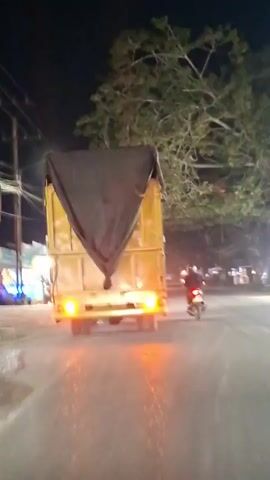 This video captures a nighttime scene on a road. A yellow truck covered with a black tarp is in the foreground, with its taillights illuminated. A motorcycle is seen in front of the truck, traveling in the same direction. The road appears to be paved, and there are streetlights casting illumination. Trees with foliage overhang the road. On the left side of the road, there are some roadside stalls or shops with lights on. The overall atmosphere suggests a rural or suburban setting at night.
