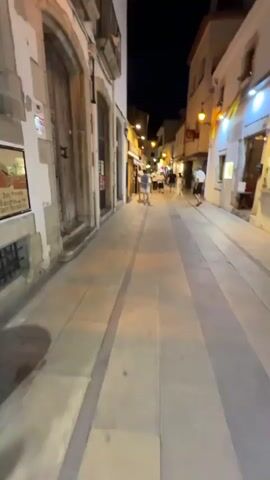 The video begins with a first-person perspective looking down a narrow, European-style street at night. The street is paved with large, square tiles, and the buildings on either side are illuminated by streetlights and interior lights from shops. The buildings are made of stone and plaster, with wooden doors and balconies. Several people are walking in the distance. As the video progresses, the camera turns to focus on a woman in a red baseball cap and blue tank top. She is holding an ice cream cone and smiling directly at the camera. She appears to be taking a selfie. Behind her, two other women are browsing merchandise displayed outside a shop. The street continues to be lit and appears to be a lively area at night.