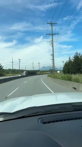 A white vehicle is driving down a highway on a sunny day with a blue sky and scattered clouds. Power lines stretch across the sky, supported by utility poles that line the side of the road. In the distance, mountains are visible. The road has multiple lanes, and other vehicles, including a truck, can be seen in the distance. The landscape is a mix of grass, trees, and what appears to be wild vegetation. The camera is positioned inside the vehicle, looking out through the windshield, showing the dashboard in the foreground.