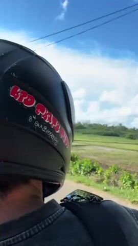 The video shows a first-person perspective of someone riding a motorcycle. The rider is wearing a black helmet with red writing. The motorcycle has a rearview mirror reflecting the rider. The scenery is rural, with green fields and trees. There are power lines and poles visible. The sky is blue with white clouds. The fields appear to be agricultural, possibly rice paddies, with some areas of water and tilled soil. In one shot, a dark object is visible hanging from what appears to be a power line.