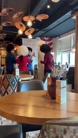 The media file depicts an indoor scene, likely a restaurant, with a focus on a group of people dressed in traditional attire. Four individuals stand in the center, two of whom are wearing large, fluffy white hats, and one person is wearing a tall, white, fur-lined garment. One person in the group is wearing a red dress, and another a blue outfit. They appear to be interacting, possibly posing for photos or engaging in a performance.

The setting is decorated with various objects. Hanging from the ceiling are lights with bulbous shades that resemble organic shapes, possibly made of wood or clay. There are also decorative elements on the walls and hanging fabrics. In the foreground, a round wooden table is set with chairs upholstered in a patterned fabric. On the table, there's a vase with dried wheat stalks and a menu or information stand. Behind the main group, another table is visible with a few people seated, one of whom is holding a phone, suggesting a public gathering or event. The lighting suggests it is daytime, possibly late afternoon.

On the menu stand, there is visible text that includes the number "15" and what appears to be a logo or emblem. The overall ambiance suggests a cultural event or a special occasion taking place in a restaurant. The presence of traditional clothing points towards a celebration of local culture or heritage. The facial expressions and body language of the individuals in the foreground are not clearly discernible due to the hats and distance. However, the atmosphere seems lively and celebratory. The location context is Monchegorsk, Russia.