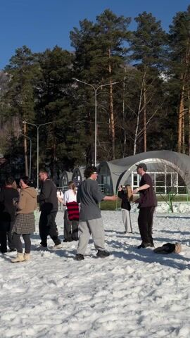 The video shows a crowd of people standing in the snow and watching a live performance on an outdoor stage. The stage is decorated with bright turquoise and green lights, with a banner that reads "МАНЖЕРОК" and the logo of "ЗВУК". A DJ and a vocalist are performing on stage. The crowd is made up of diverse individuals in winter clothing, some of whom are filming the performance with their phones. The background consists of a snow-covered ground, tall evergreen trees, a clear blue sky, and a distant mountain. The overall atmosphere suggests an outdoor winter music event.