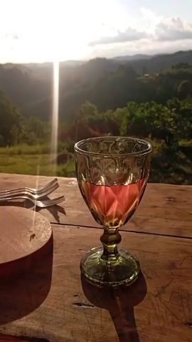 The video shows a close-up of a wooden table with a glass of pink liquid, a round wooden cutting board, and three forks. In the background, there is a lush green landscape of rolling hills and trees bathed in the warm glow of the setting sun. The sky is partly cloudy, and a strong sunbeam cuts through the clouds. The scene appears to be set outdoors, likely on a balcony or deck overlooking a valley. There are no people visible in the video.