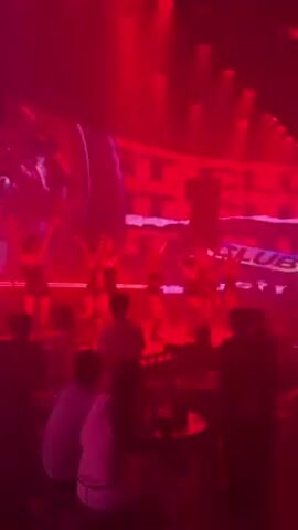 A group of women in revealing costumes performs on a stage in a nightclub. The women are wearing short shorts and tops or bodysuits. The lighting is primarily red, with some white spotlights. The audience sits at tables in front of the stage, watching the performance. There's a large screen behind the performers. The nightclub is crowded, and the atmosphere is lively.
