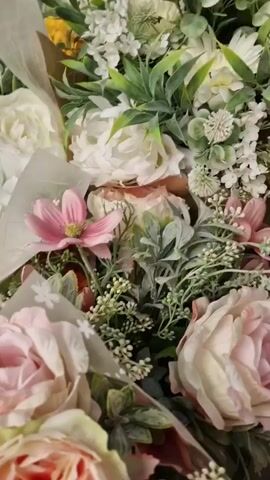 The video is a close-up, top-down shot of a bouquet of artificial flowers. The bouquet features various types of flowers in shades of pink, white, and peach. Among the flowers are several large, ruffled roses and smaller, star-shaped pink flowers. Green foliage and small white buds are interspersed throughout the arrangement. The flowers are wrapped in translucent paper with a subtle star pattern. The lighting is soft and natural, highlighting the textures and colors of the artificial blooms. The video is shot with a shallow depth of field, blurring the background and focusing attention on the central elements of the bouquet. The camera slowly pans across the arrangement.