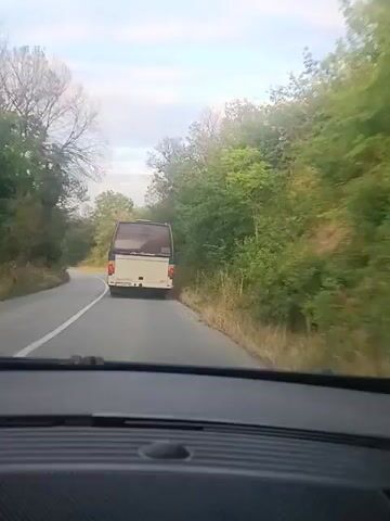 The video captures a view from inside a vehicle, looking ahead at a road curving to the left. A white and blue bus is in front, traveling in the same direction. The road is paved and has a white line marking its edge. On either side of the road, there are trees and foliage, indicating a natural, possibly rural, environment. The sky above is partly cloudy, with soft lighting suggesting it might be morning or late afternoon. The camera appears to be positioned behind the dashboard of the vehicle, with the upper edge of the dashboard visible in the foreground. The bus is the primary focus of the moving elements in the scene, as it drives away from the viewer.