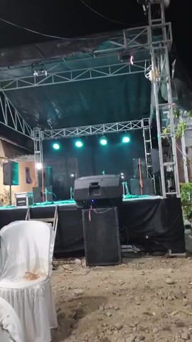 A temporary outdoor stage is set up at night, ready for an event. The stage is constructed with a metal truss system supporting a black tarp roof. Blue lights illuminate the stage from above, casting a glow on the equipment below. Several speakers, including large subwoofers and smaller monitors, are positioned on and around the stage. A black curtain drapes the front of the stage. In the foreground, several chairs are covered with white cloths, suggesting the seating area for an audience. The ground appears to be dirt and gravel. The overall scene is one of preparation for a musical performance or other outdoor gathering.