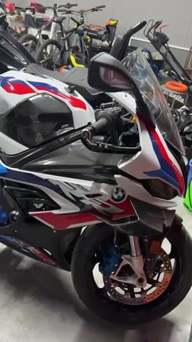 The video shows a garage with two motorcycles parked next to each other. The first motorcycle, closest to the camera, is a BMW sport bike with white, blue, and red detailing and carbon fiber body parts.  Next to it is a larger black motorcycle with a fairing. In the background, there are multiple bicycles and various garage tools and equipment, including a lawnmower. The garage floor is gray and reflective. The overall lighting in the garage appears adequate.