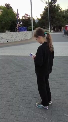 A young girl with a ponytail, wearing a black sweater and black pants, stands facing away from the camera, looking at a smartphone in her hands. She is on a paved walkway made of interlocking bricks. In the background, there are trees, a stone wall, and a large sign with purple lettering that appears to spell out "CHANCE." Further in the distance, there are stairs leading up to a plaza with buildings and a monument. The sky is clear and bright, suggesting it is daytime.