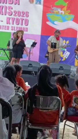 A group of performers is on a stage in front of an audience. The stage backdrop features a banner with text that includes "KIS 2026 - KYOTO - KABUPATEN SLEMAN" and a colorful graphic of the Earth with animals. On the left side of the stage, a woman in a black dress and hat is singing into a microphone, with a keyboard and a man in a suit behind her. In the center right, a man wearing a hat and glasses is playing an acoustic guitar and singing. The audience members are seated in chairs, many of whom are wearing orange shirts and some are wearing hijabs. The environment appears to be outdoors or in a large open space, possibly a community event or festival.