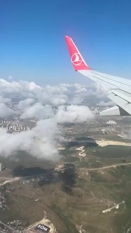An aerial view from an airplane window shows a clear blue sky with scattered white clouds. The wing of a Turkish Airlines plane, with its distinctive red logo, is visible in the foreground. Below, a cityscape with buildings can be seen through gaps in the clouds, along with green hills and a partially visible road. The lighting suggests it is daytime.